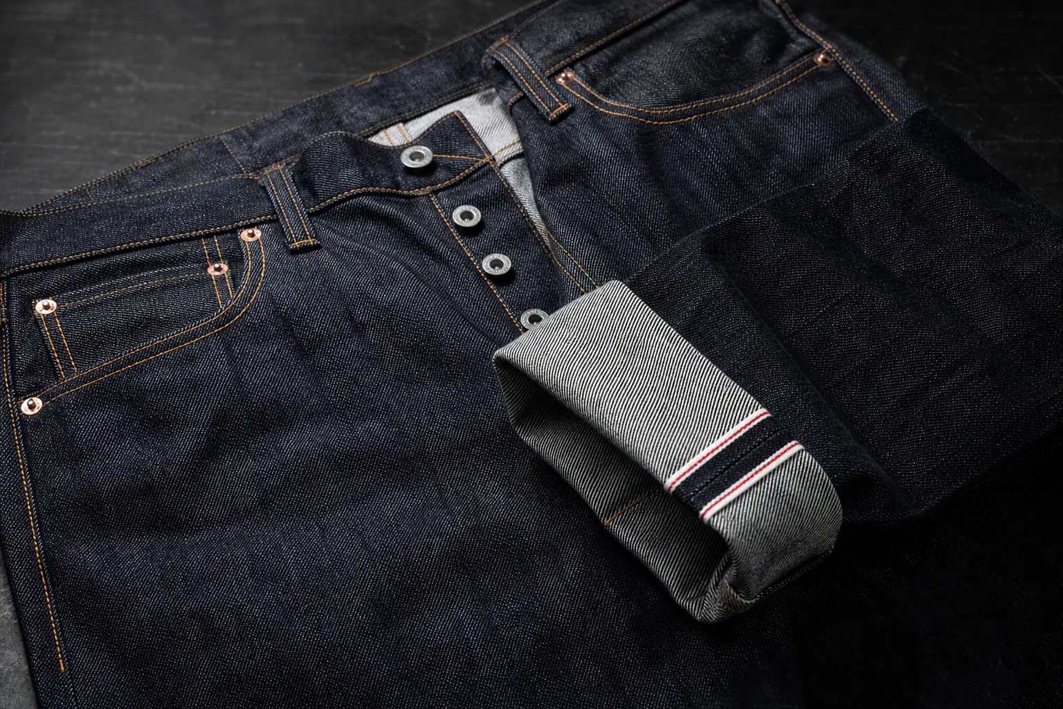 Raw selvedge denim showing its cuff edge, button fly, rivets, and dense twill